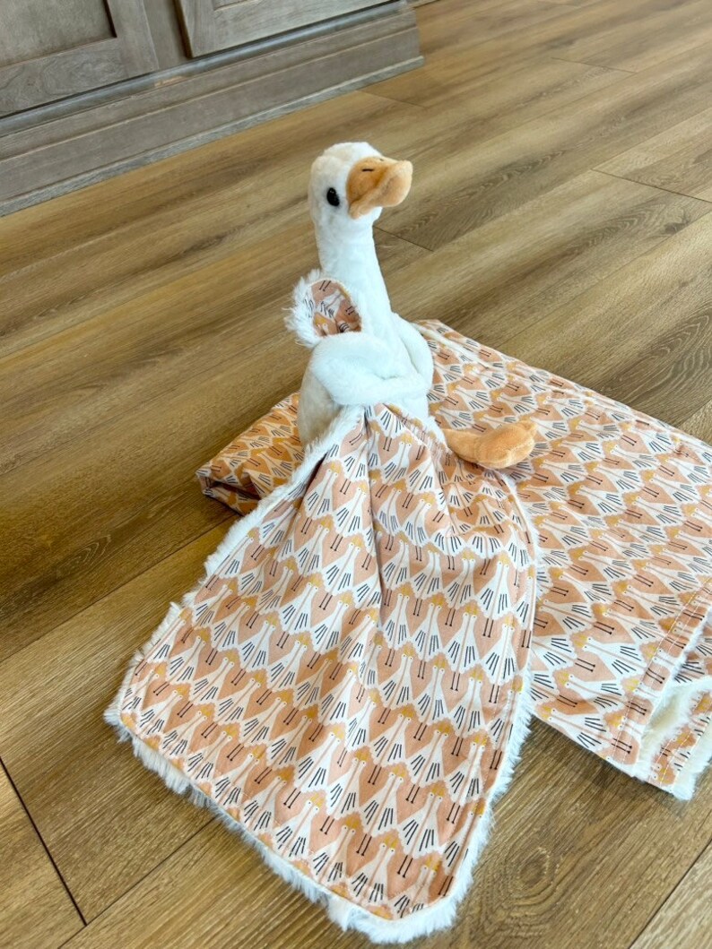 May include: A white plush goose toy is lying on a brown and white patterned baby blanket. The blanket has a soft, fuzzy texture and features a repeating pattern of small geese.