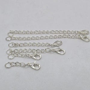 May include: A collection of five silver-toned metal necklace extender chains of varying lengths. Each chain features a circular jump ring at one end and a lobster claw clasp at the other end, designed to increase the length of jewelry pieces.