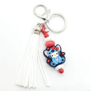 May include: A white tassel keychain with a blue and black cartoon character charm. The charm has red hearts and a red bow. The keychain also has red and white beads.