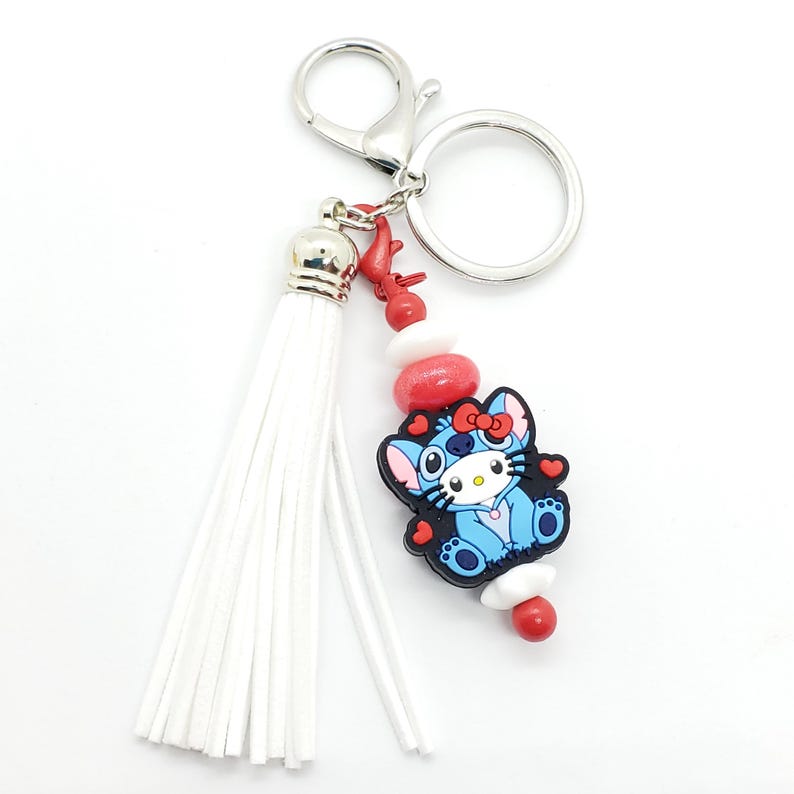 May include: A white tassel keychain with a blue and black cartoon character charm. The charm has red hearts and a red bow. The keychain also has red and white beads.