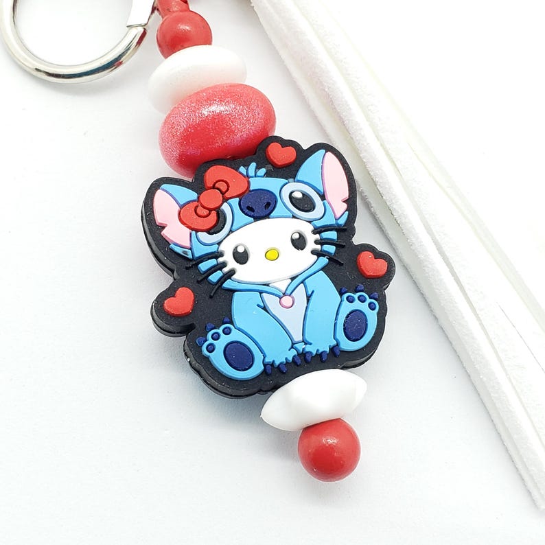 May include: A blue Stitch character wearing a Hello Kitty costume with red hearts and a white tassel keychain. The keychain has red and white beads.