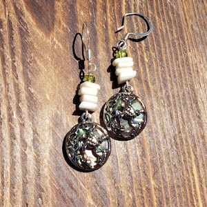 May include: Silver-toned dangle earrings with a round, iridescent pendant featuring a turtle design. The earrings have white, rectangular beads and green beads. The earrings are displayed on a wooden surface.
