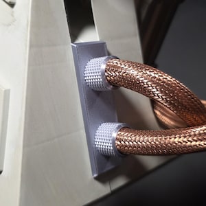 May include: Close-up of a computer component featuring copper-colored braided tubing connected to a gray rectangular plate with silver-colored, textured connectors. The component is set against a light beige background.