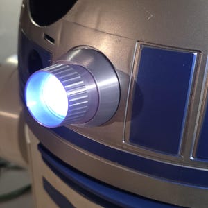 May include: A silver and blue robotic head featuring a prominent circular lens that emits a bright white light. The structure includes a cylindrical housing with vertical ridges surrounding the light source, set against a metallic dome surface.