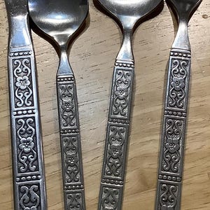 Könnte beinhalten: Ein Satz Vintage-Besteck, bestehend aus einem Messer, zwei Löffeln und einer Gabel. Die Edelstahlutensilien weisen verzierte Blumenmuster auf den Griffen auf. Das Besteck ist auf einer Holzoberfläche angeordnet.