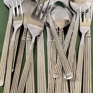 May include: A collection of silver-toned cutlery, including forks, knives, and spoons, with a textured design on the handles. The cutlery is arranged in a pile on a green surface.