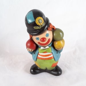 May include: A ceramic clown figurine with red hair, a black hat with a blue and yellow flower, and a red, green, and yellow ball in each hand.