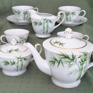 Może przedstawiać: Biały porcelanowy zestaw do herbaty z wzorem bambusa. Zestaw zawiera czajnik, dzbanek na śmietanę, cukiernicę i cztery filiżanki do herbaty z podstawkami.