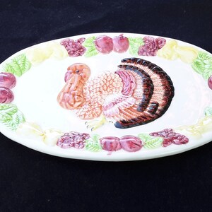 May include: White oval ceramic serving platter featuring a raised, painted image of a turkey surrounded by a border of green leaves, red grapes, and yellow pears.