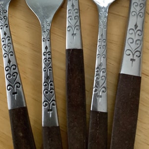 Può includere: Set di cinque posate argentate con manici in legno marrone. Le posate includono una forchetta, un coltello e tre cucchiai. I manici hanno un motivo floreale decorativo.