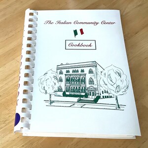 Könnte beinhalten: Ein spiralgebundenes Kochbuch mit dem Titel The Italian Community Center Cookbook mit weißem Einband, das eine grüne Strichzeichnung eines Gebäudes, zwei Bäume und ein kleines Symbol der italienischen Flagge zeigt.