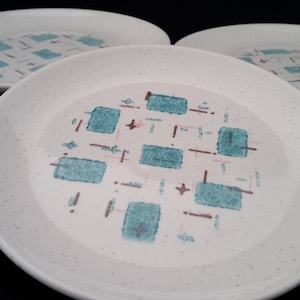 Op de afbeelding: Set van vier witte keramische dinerborden met een geometrisch patroon van tealblauwe en bruine vierkanten en lijnen op een gespikkelde achtergrond.