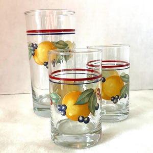 May include: Three clear glass tumblers of varying heights featuring a painted design of yellow oranges, green leaves, and small dark blue berries. Each glass is decorated with a horizontal red and blue stripe encircling the upper rim area.