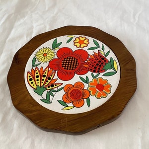 Op de afbeelding: Een houten onderzetter met een witte keramische tegel met een kleurrijk bloemenontwerp. De tegel heeft rode, oranje, gele en zwarte bloemen met groene bladeren.