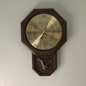 Puede incluir: Un barómetro con marco de madera con una cara dorada y un diseño de águila. La cara tiene un dial con los números del 1 al 31 y las palabras "Lluvia" y "Bueno" con una aguja que apunta a "Cambio".