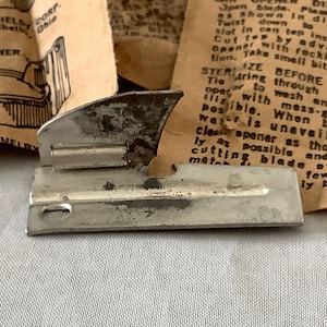 Puede incluir: Un abrelatas de plata con una cuchilla afilada y un pequeño mango. El abrelatas está diseñado para cortar la parte superior de una lata. Las instrucciones para usar el abrelatas están impresas en un trozo de papel detrás del abrelatas.