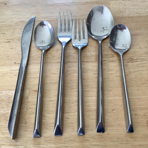 Pode incluir: Um conjunto de seis talheres de aço inoxidável, incluindo uma faca, dois garfos e três colheres. Os utensílios têm um design simples e moderno com cabos longos e finos e um acabamento polido. Os talheres estão dispostos sobre uma superfície de madeira clara.