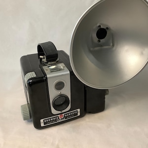 May include: A black and silver Brownie Hawkeye Flash camera with a detachable flash unit. The camera has a red and white logo that says "Brownie Hawkeye Flash".