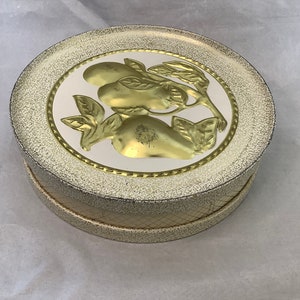 Puede incluir: Una caja redonda dorada vintage con tapa. La tapa presenta un diseño dorado en relieve de frutas y hojas sobre un fondo blanco. La caja tiene un borde dorado texturizado y moteado y un lateral estampado.