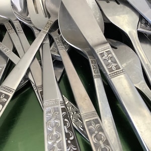 May include: A selection of stainless steel cutlery, including spoons, forks, and knives. The handles have an embossed floral pattern. The cutlery is piled together, with the shiny metal reflecting light.
