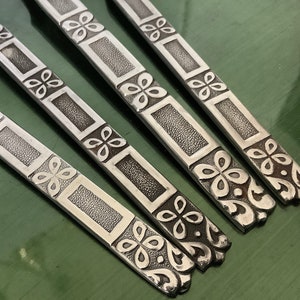 以下が含まれることがあります： 装飾的な花の模様が施された銀色のフラットウェア4点。模様は四つ葉のクローバーを繰り返したデザインです。