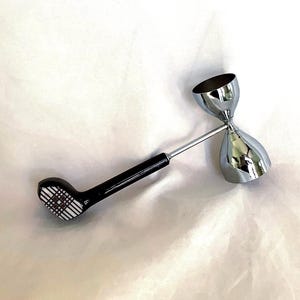 Könnte beinhalten: Ein Cocktail-Jigger aus Edelstahl mit schwarzem Griff. Der Griff ist wie ein Golfschläger geformt, mit einem Karomuster auf dem Schlägerkopf. Der Jigger hat zwei Messbecher, die durch einen dünnen Metallstab verbunden sind.