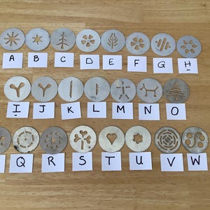May include: A set of 23 metal cookie cutters in diverse shapes, such as stars, trees, and flowers. Each cutter is labelled with a letter from A to W on a small white card. The cutters are displayed on a wooden surface.