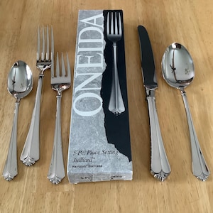 May include: A set of six stainless steel cutlery pieces, including forks, spoons, and a knife, arranged on a wooden surface. The cutlery is from the Oneida Juilliard collection and is displayed with its original packaging.