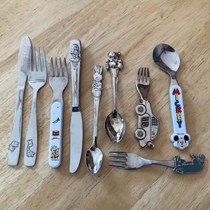 May include: A set of eight children's silverware pieces, including a knife, forks, and spoons. The handles feature various cartoon characters, including Mickey Mouse, a car, and a bear.