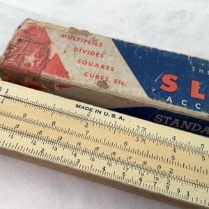 Może przedstawiać: Zabytkowa suwakowa linijka wykonana z drewna z nadrukowanymi oznaczeniami, wraz z oryginalnym kartonowym pudełkiem. Pudełko zawiera tekst wskazujący na jej zastosowanie do mnożenia, dzielenia itp. Linijka jest oznaczona "Made in U.S.A."