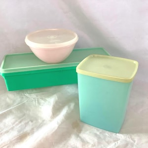 May include: A collection of three plastic food storage containers. One is a rectangular bright green box with a matching lid, one is a small round pale pink bowl with a translucent lid, and one is a tall square light blue canister with a tan lid.