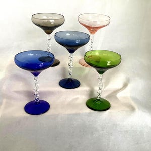 May include: Five colorful champagne coupe glasses. Each glass has a different colored bowl: blue, green, pink, gray, and blue. The stems are clear with a twisted design, and the bases match the bowl colors.