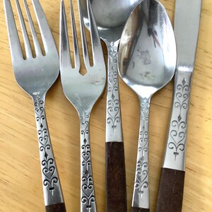 May include: A set of stainless steel cutlery including two forks, two spoons, and one knife. Each piece features a dark brown handle and a silver-toned metal neck decorated with an engraved scrollwork pattern consisting of repeating loops and diamonds.