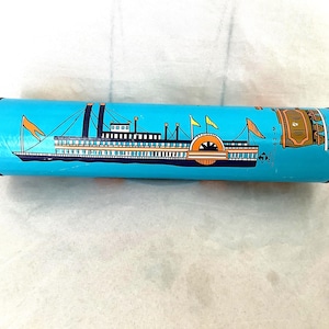 May include: A cylindrical, blue container with a black rim, featuring a detailed illustration of a steamboat with orange and yellow accents. The design includes flags and a paddle wheel. The text "63-M" is visible.