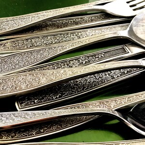 May include: Close-up of silver cutlery, including spoons and a fork. The handles have a detailed floral pattern. The silverware is arranged on a green surface, contrasting with the metallic sheen. The image shows a close-up of the silverware.
