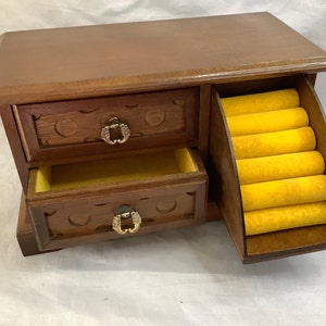 Op de afbeelding: Een houten juwelendoos met twee laden en een ringhouder. De doos is bruin met een lichte houtnerf en heeft messing accenten. De ringhouder is bekleed met geel vilt.