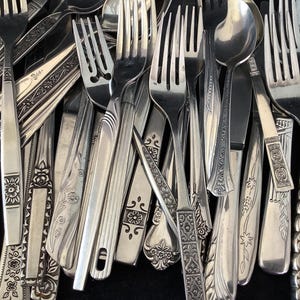 Op de afbeelding: Een close-up van een verzameling vintage zilverwerk, waaronder vorken, lepels en messen. Het zilveren bestek heeft ingewikkelde handvatontwerpen. De afbeelding toont een verscheidenheid aan patronen en texturen.