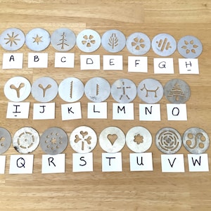 May include: A collection of twenty-three circular metal cookie press discs, each featuring a unique cutout design. The discs are arranged in three rows, with each disc labelled by a white paper square marked with a letter from A through W.