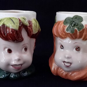 Könnte beinhalten: Zwei Keramikbecher in Form von Gesichtern. Der Becher links hat braunes Haar und grüne Blätter um den Kopf. Der Becher rechts hat orangefarbenes Haar und ein grünes Kleeblatt auf dem Kopf.