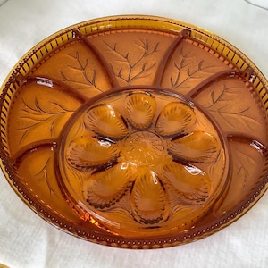 Puede incluir: Fuente de vidrio color ámbar con un cuenco central y seis compartimentos circundantes. La fuente presenta un diseño de hojas y un borde decorativo. Una pieza vintage para servir aperitivos.