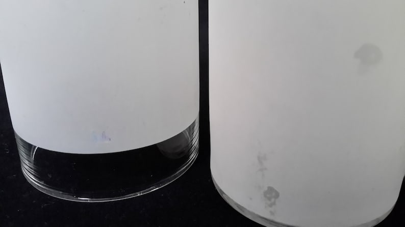 May include: Two white frosted glass cylinder vases with a clear glass base. The vase on the right has a few small dark spots on the frosted surface.