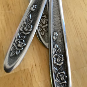 May include: Close-up of several silver-colored utensils with a floral rose design. The rose pattern is embossed on the handles of the utensils. The utensils are arranged on a light brown wooden surface.