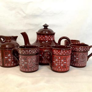 Könnte beinhalten: Ein komplettes braunes Teeservice mit passendem Sahnekännchen, Zuckerdose, Teekanne und Tassen. Das Set hat ein rotes und weißes Paisley-Design. Die Teekanne hat einen Deckel und einen gebogenen Ausguss. Die Tassen haben Henkel.