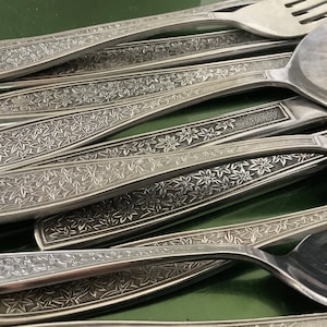 Puede incluir: Primer plano de una colección de cubiertos de acero inoxidable. Los cubiertos incluyen cucharas y tenedores. Los mangos están decorados con un patrón floral repetitivo. Los cubiertos están dispuestos sobre una superficie verde.