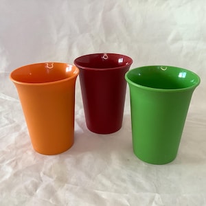 Könnte beinhalten: Drei bunte Plastikbecher in Orange, Rot und Grün. Die Becher haben einen leicht ausgestellten Rand und eine glatte, glänzende Oberfläche. Sie sind auf einer weißen, strukturierten Oberfläche angeordnet.