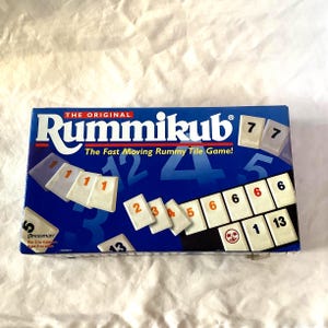 May include: A blue box of the game Rummikub, with white tiles displaying numbers. The box has the game's name in white and red text, and the phrase "The Fast Moving Rummy Tile Game!" is also visible.