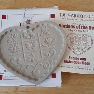 Puede incluir: Molde para galletas de cerámica blanca en forma de corazón con un diseño en relieve de flores, mariposas y hojas. El molde está en una caja blanca con el texto "The Pampered Chef" y "Gardens of the Heart Clay Cookie Mold".