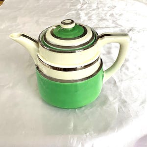 Op de afbeelding: Een vintage keramische theepot met een groene basis, een crèmekleurig middengedeelte en een groen en crèmekleurig gestreept deksel. Zilverkleurige randen accentueren het ontwerp. De theepot heeft een gebogen tuit en een grote handgreep.