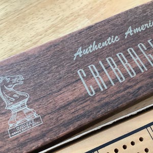 Puede incluir: Tablero de cribbage de madera con el texto "Authentic American Cribbage Board" y un diseño de pieza de ajedrez de caballero. El tablero tiene un patrón de vetas de madera y la marca "Druke" es visible.