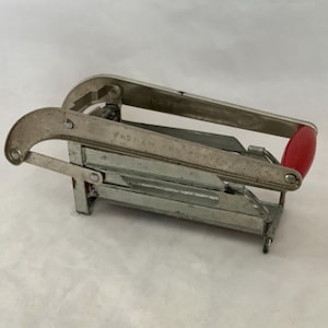 May include: A vintage, silver-toned metal can opener with a red handle. The can opener has a lever and a curved blade. The handle is a solid, rounded shape. The can opener is designed to open cans.
